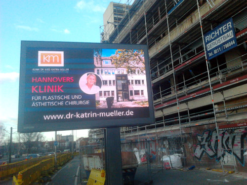Werbetafel mit wechselnden Inhalten vor der verrotteten Fassade der Ruine des Ihmezentrums: km -- Hannovers Klinik für plastische und ästhetische Chirurgie -- www.dr-katrin-mueller.de